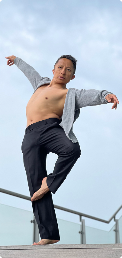 Kwok One gracefully poses in a dance stance on one foot, arms extended. They wear a grey cardigan, black pants, and the background shows a cloudy sky and railing.