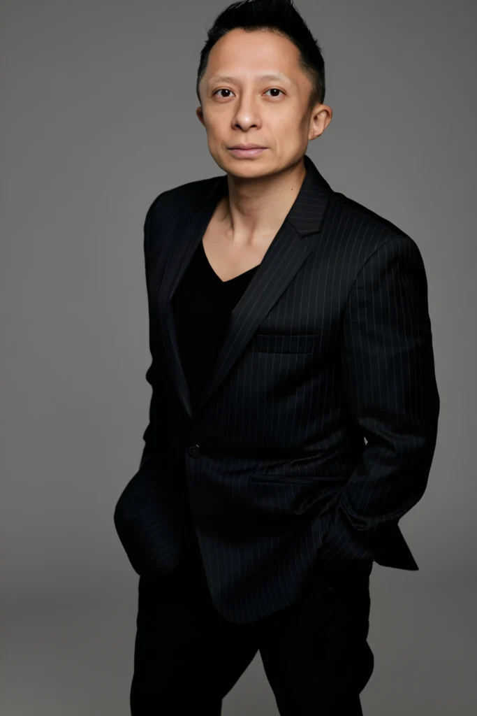 Professional portrait of Kwok One in a dark pinstripe suit against a grey background – corporate headshot with confident expression and formal attire.
